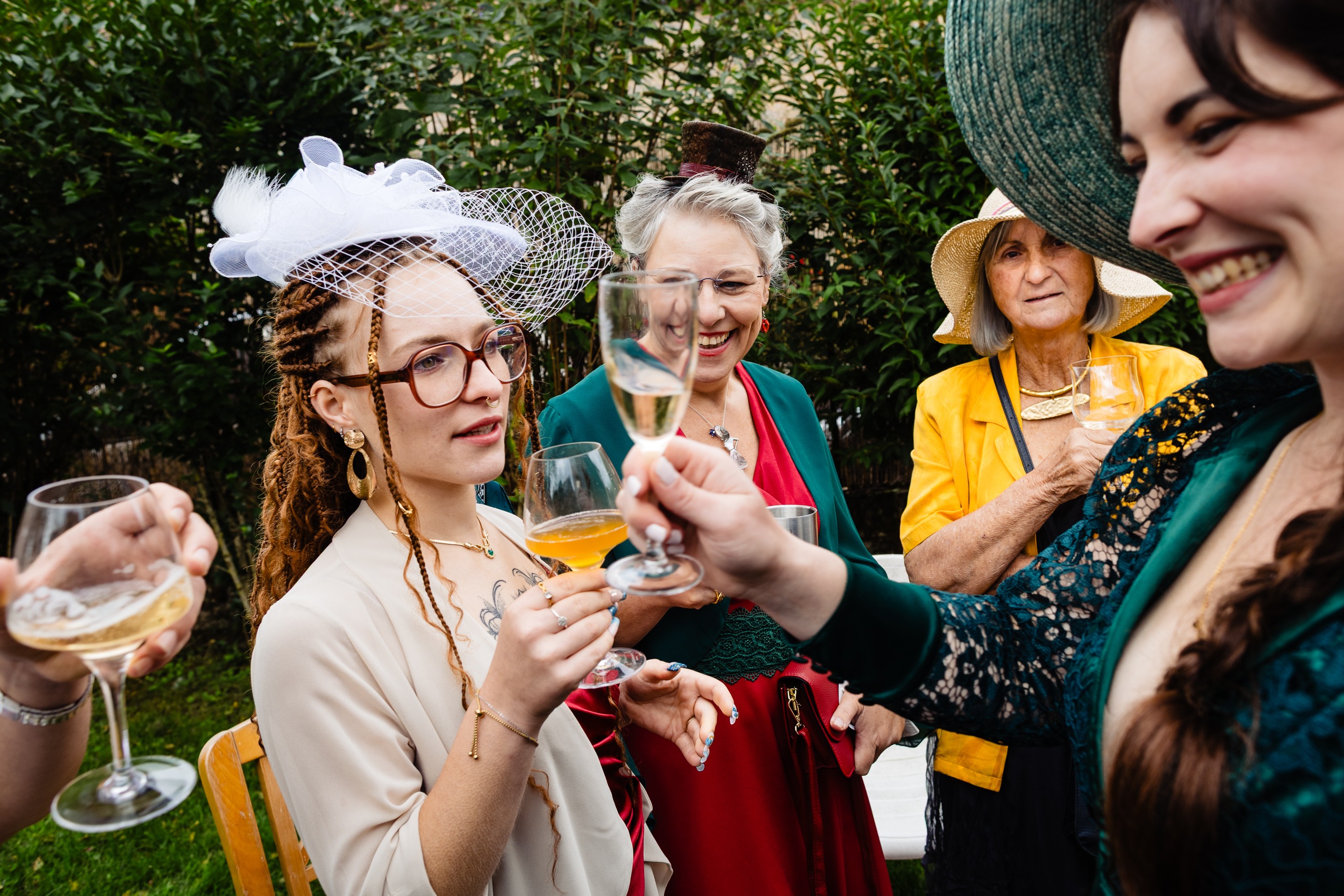 Un groupe de femmes vêtues de vêtements colorés et formels et de chapeaux tintent des verres et sourient lors d'une réunion en plein air.