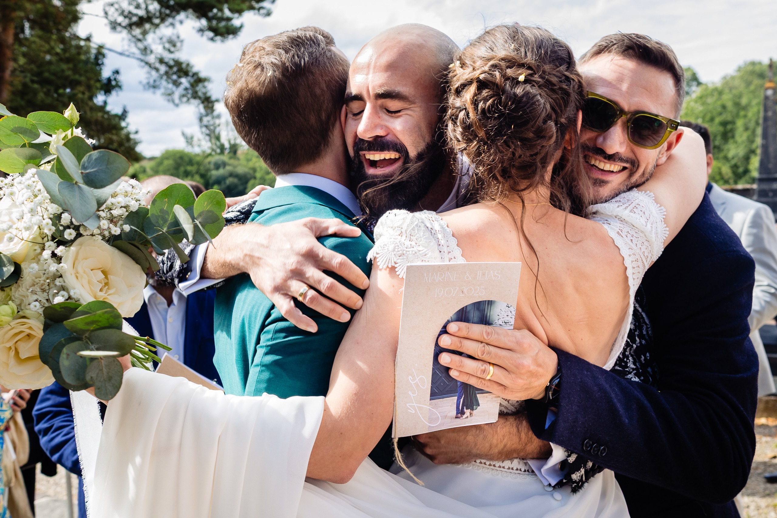 Quatre personnes s'étreignent joyeusement en plein air lors d'un mariage ; l'une tient un bouquet de roses blanches et de verdure, l'autre un programme de mariage.