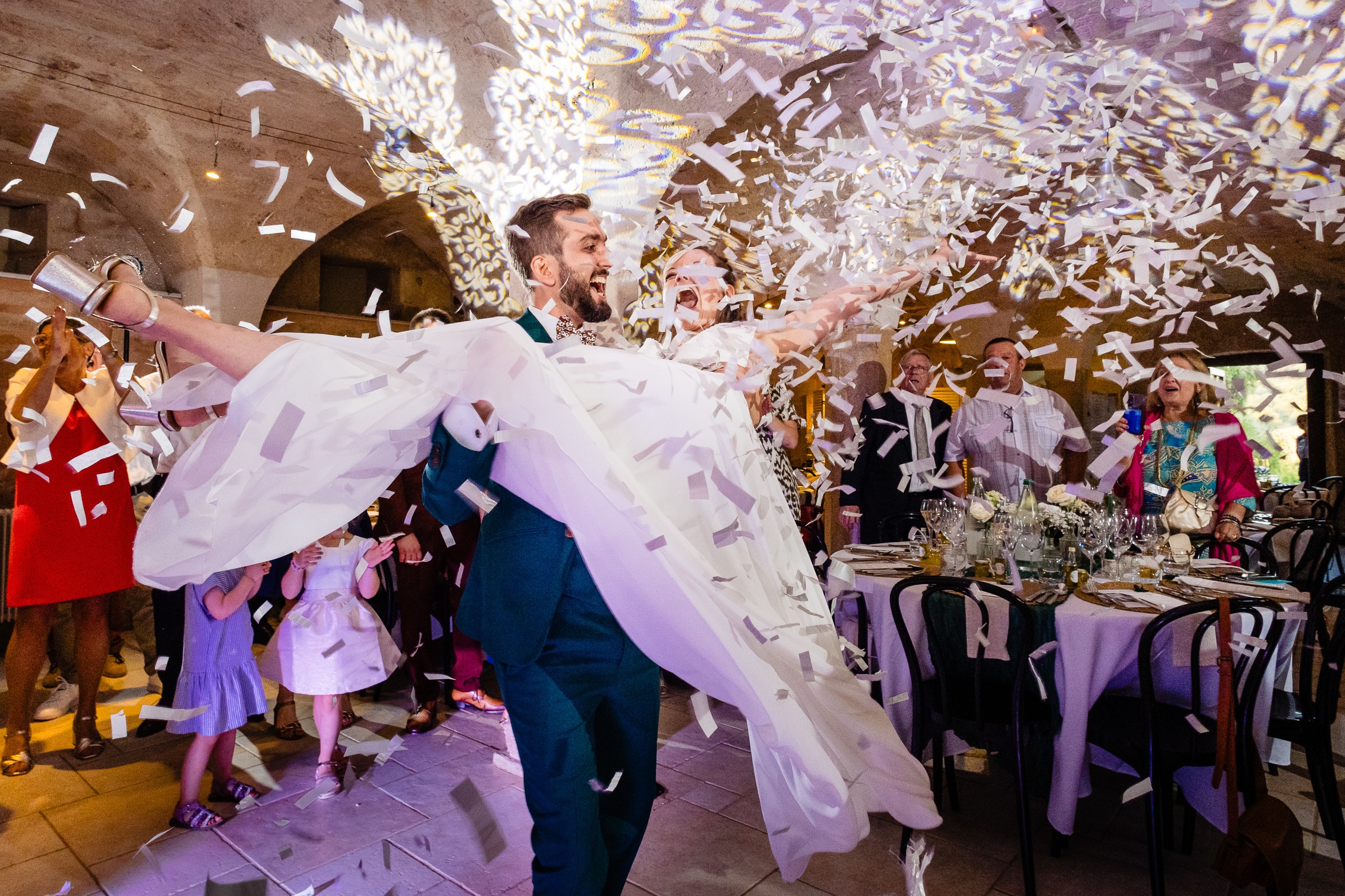 Un marié soulève une mariée tandis que des confettis tombent autour d'eux lors d'une réception de mariage, entourés d'invités en liesse et de tables décorées.