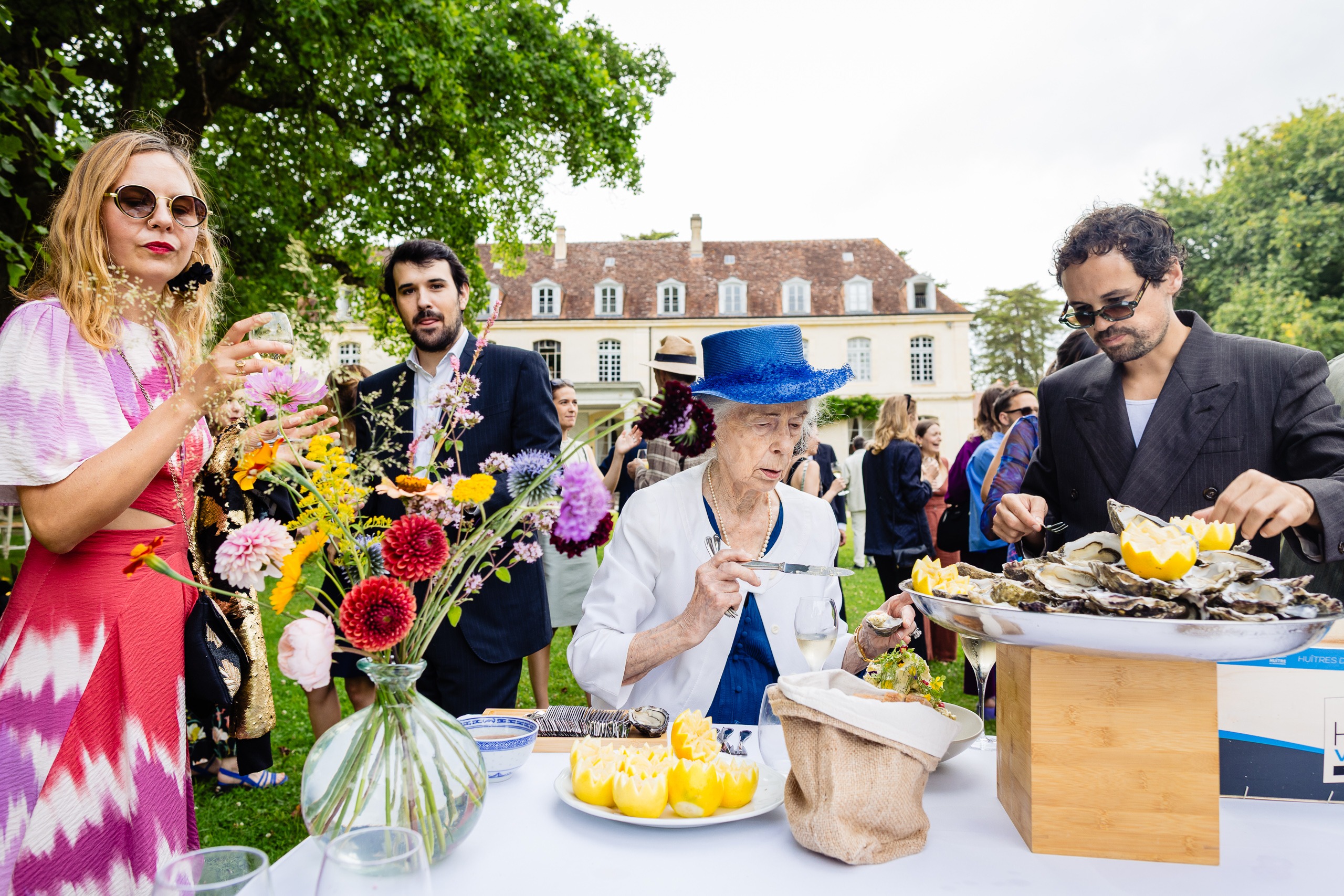 Un groupe de personnes, dont une femme âgée coiffée d'un chapeau bleu, se réunit autour d'une table en plein air avec des fleurs et un plateau d'huîtres lors d'un événement dans un jardin.