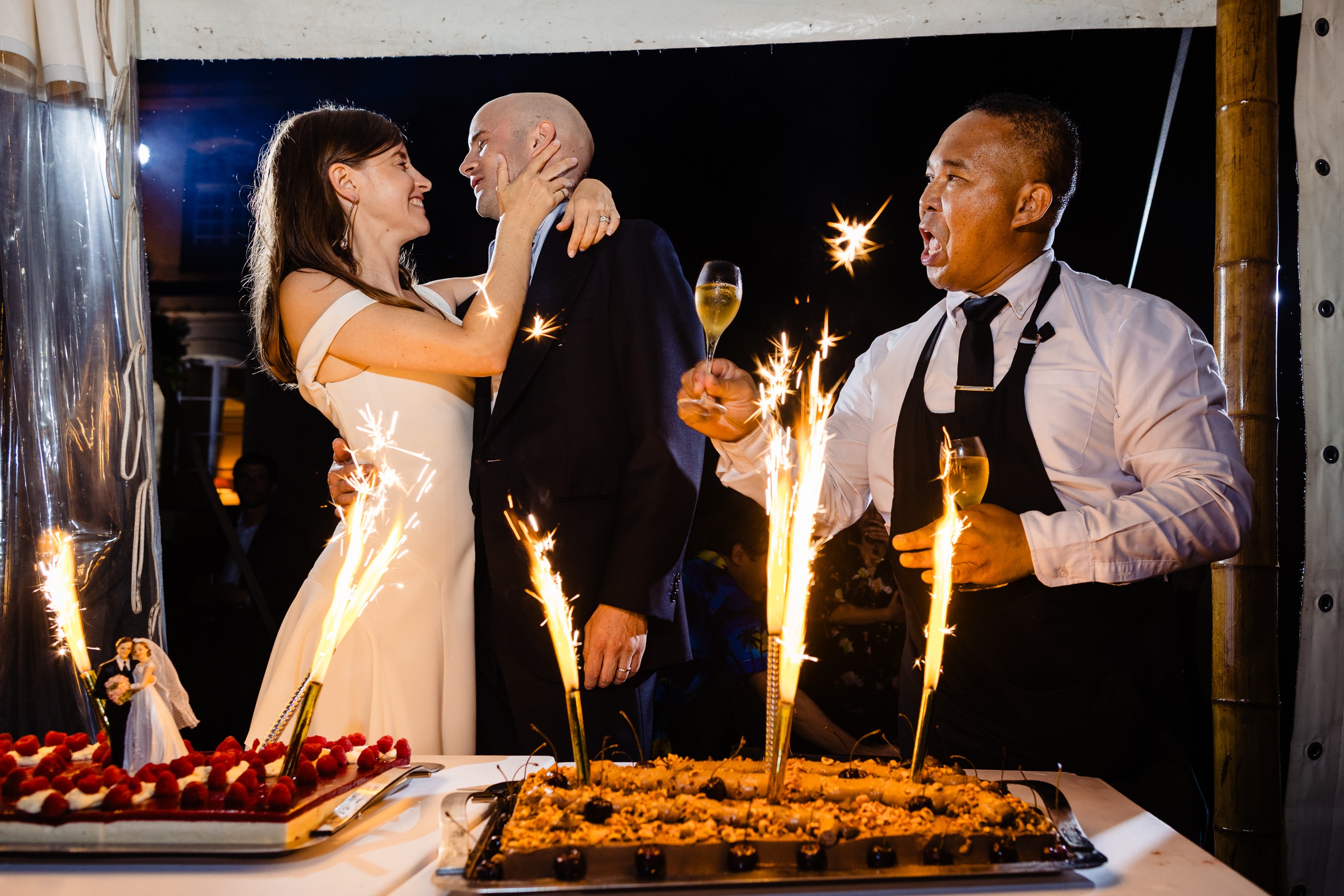 Des mariés se sourient près de gâteaux avec des cierges magiques, tandis qu'un homme en tablier fête l'événement avec une coupe de champagne.