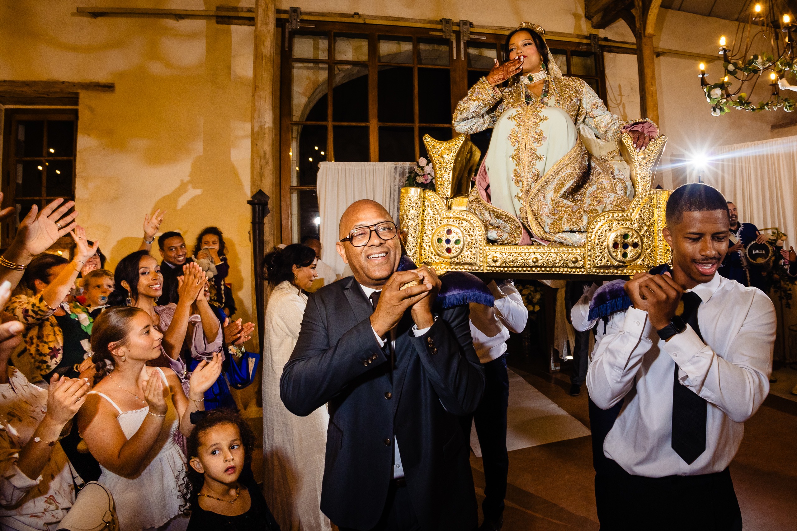 Une femme vêtue d'une tenue raffinée est assise sur une chaise décorative portée par deux hommes lors d'une fête en intérieur, tandis que les invités applaudissent et sourient.