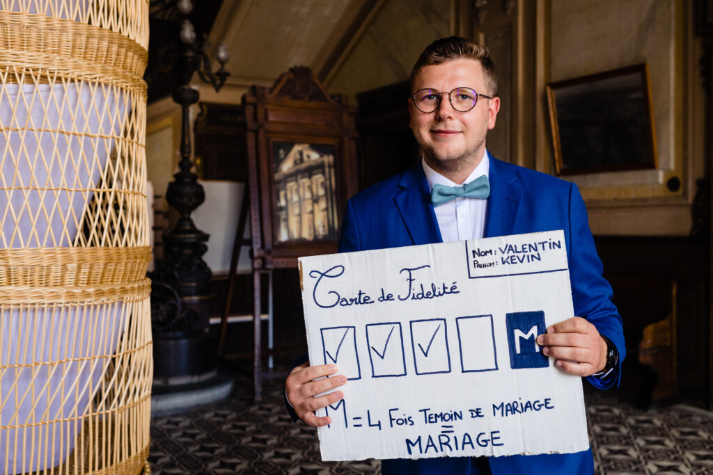 Un homme en costume bleu se tient à l'intérieur de la Ferme Sainte-Anne, près de Langres, et tient une pancarte faite à la main en français sur le fait d'être témoin de mariage à plusieurs reprises, avec des cases à cocher et des détails écrits à la main - parfait pour une cérémonie laïque.