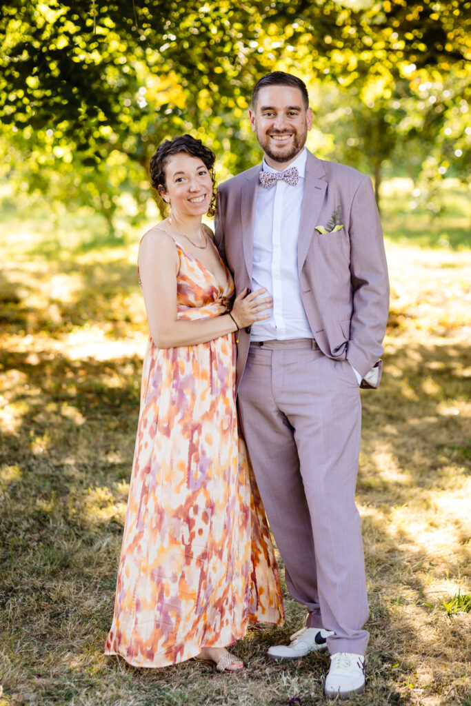 Amélie Kevin se tient en plein air sur l'herbe au Mariage Ferme Saint-Anne, à Langres ; l'une porte une robe sans manches à motifs, l'autre un costume mauve avec un nœud papillon et des baskets. La lumière du soleil filtre à travers les arbres derrière eux.