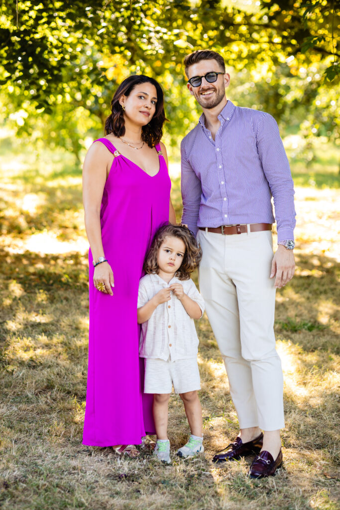 À la Ferme Saint-Anne de Langres, Amélie et Kevin posent avec un jeune enfant sur l'herbe, sous les arbres - une femme en robe rose vif, un homme en chemise rayée et pantalon clair - capturant un moment spécial de leur mariage.