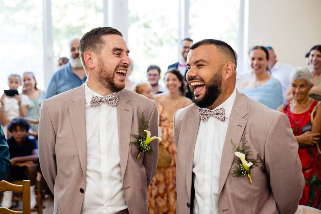 Deux hommes en costume clair et nœud papillon assortis se tiennent côte à côte, arborant un large sourire lors du mariage d'Amélie et Kevin, entourés d'un groupe joyeux dans la salle lumineuse de la Ferme Saint-Anne de Langres.