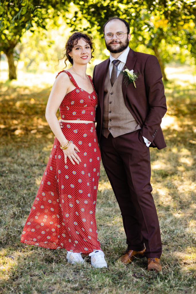 Amélie Kevin pose en plein air au Mariage Ferme Saint-Anne à Langres - une femme vêtue d'une robe rouge à motifs et de baskets blanches se tient à côté d'un homme vêtu d'un costume et d'un gilet marron, avec de l'herbe sous les pieds et des arbres en arrière-plan.