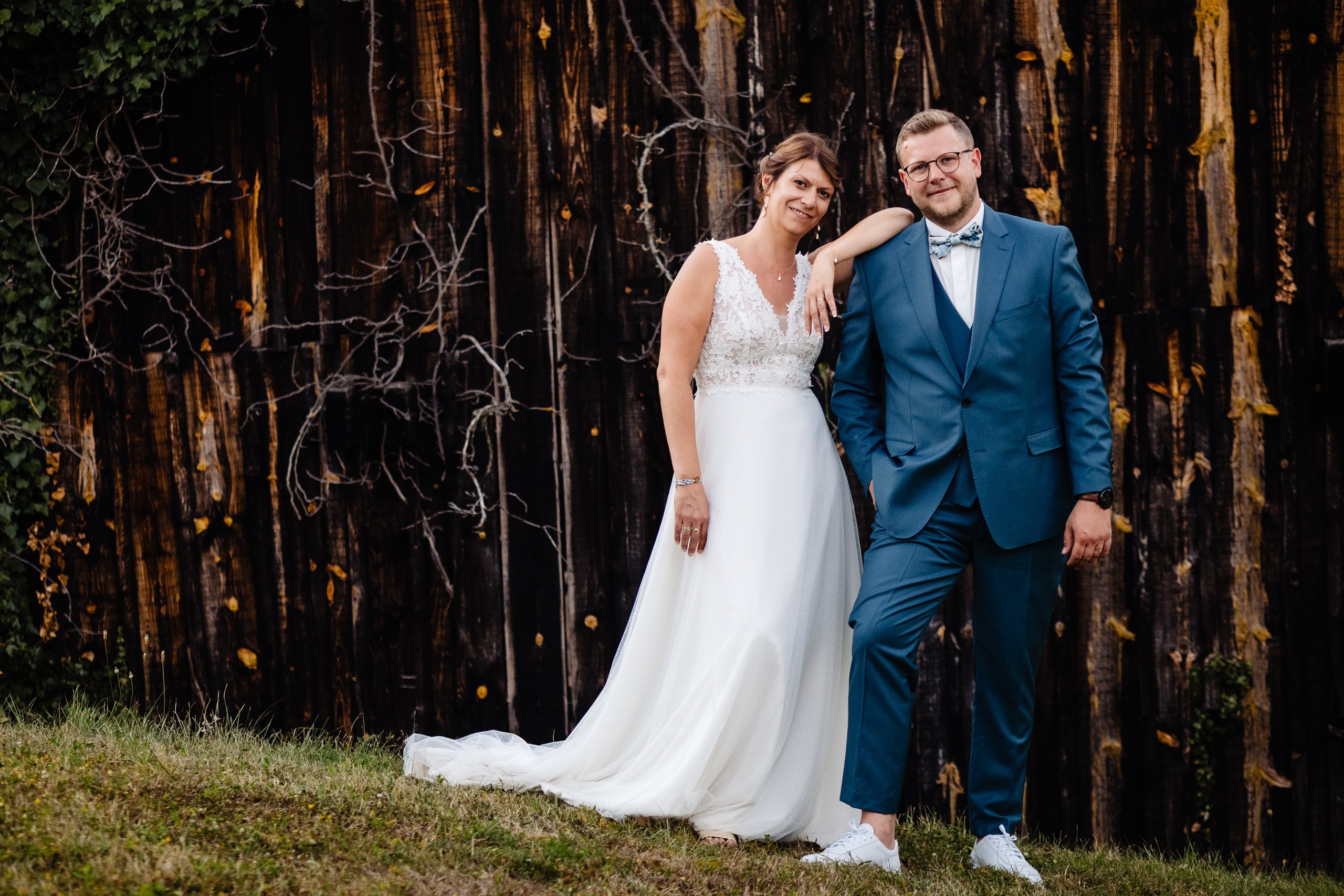Amélie & Kevin, la mariée en robe blanche et le marié en costume bleu, posent ensemble sur l'herbe devant le mur en bois rustique de la Ferme Saint-Anne de Langres.