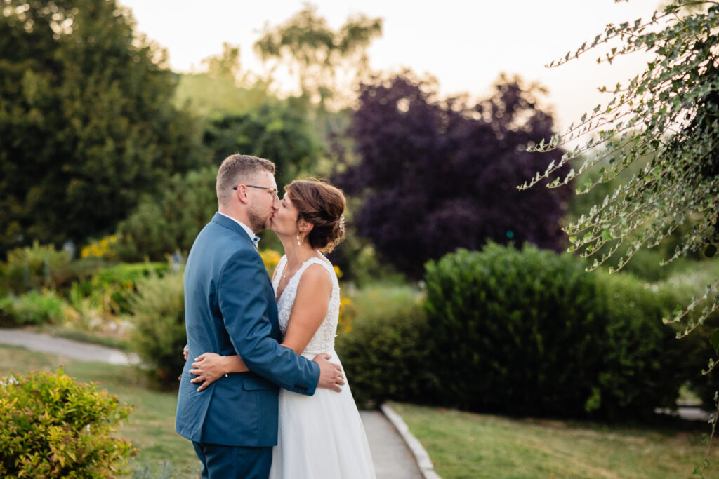 Amélie et Kevin partagent un baiser romantique dans le jardin de la Ferme Saint-Anne de Langres, entourés de verdure et d'arbres luxuriants le jour de leur mariage.