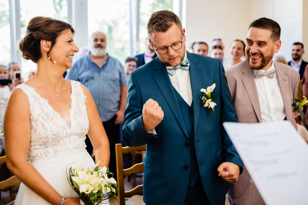 Lors du mariage d'Amélie et de Kevin à la Ferme Saint-Anne de Langres, une mariée et deux garçons d'honneur se tiennent à l'intérieur ; l'homme en costume bleu fait un poing de célébration tandis que la mariée et l'homme en beige sourient, sous le regard des invités à l'arrière-plan.