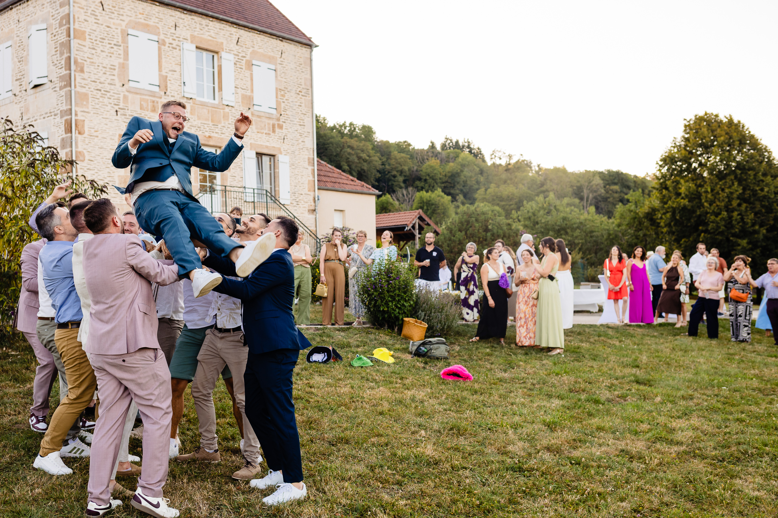 Groupe soulève Kevin en costume bleu dans les airs sur la pelouse, joie collective Ferme Sainte-Anne