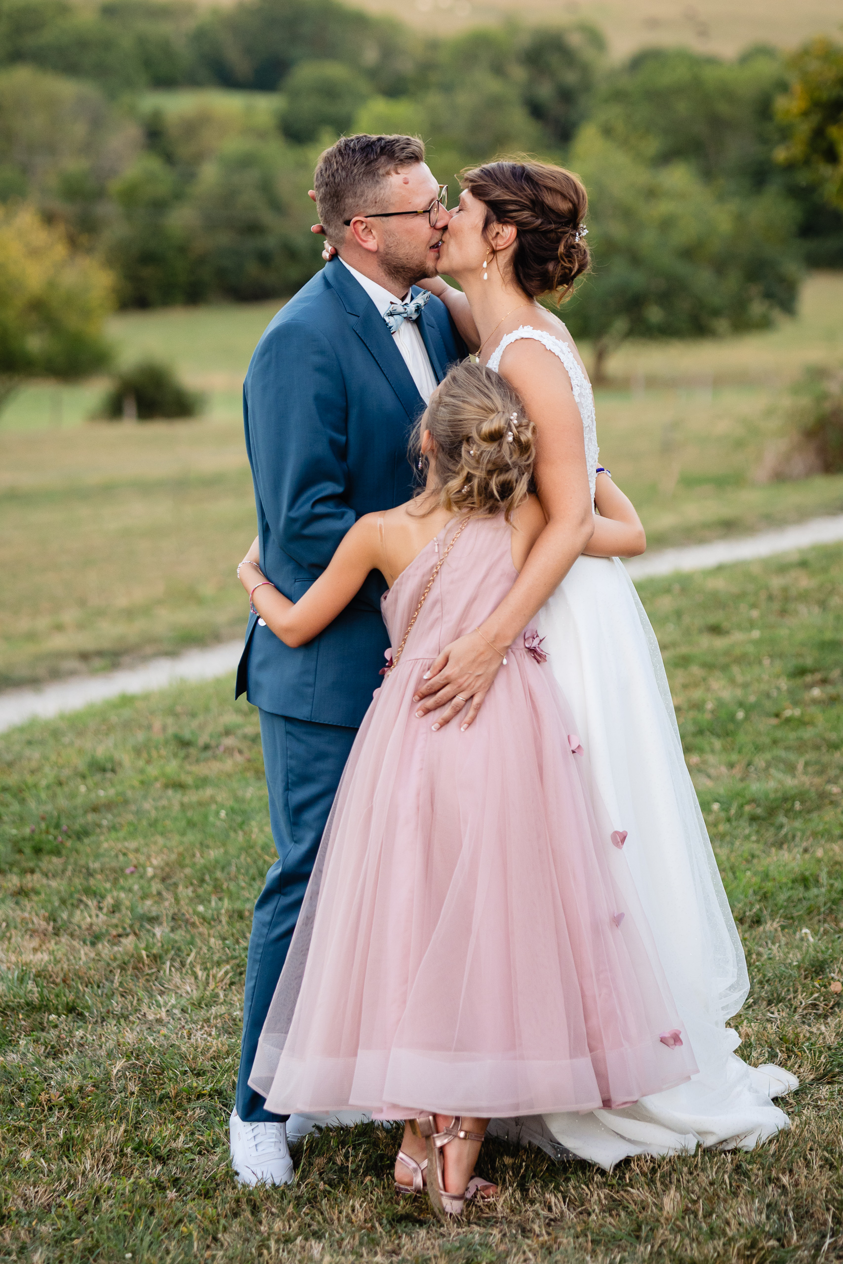 Amélie et Kevin s'embrassent en plein air lors de leur mariage, tandis qu'une jeune fille vêtue d'une robe rose les enlace par derrière sur le terrain herbeux de la Ferme Saint-Anne de Langres.