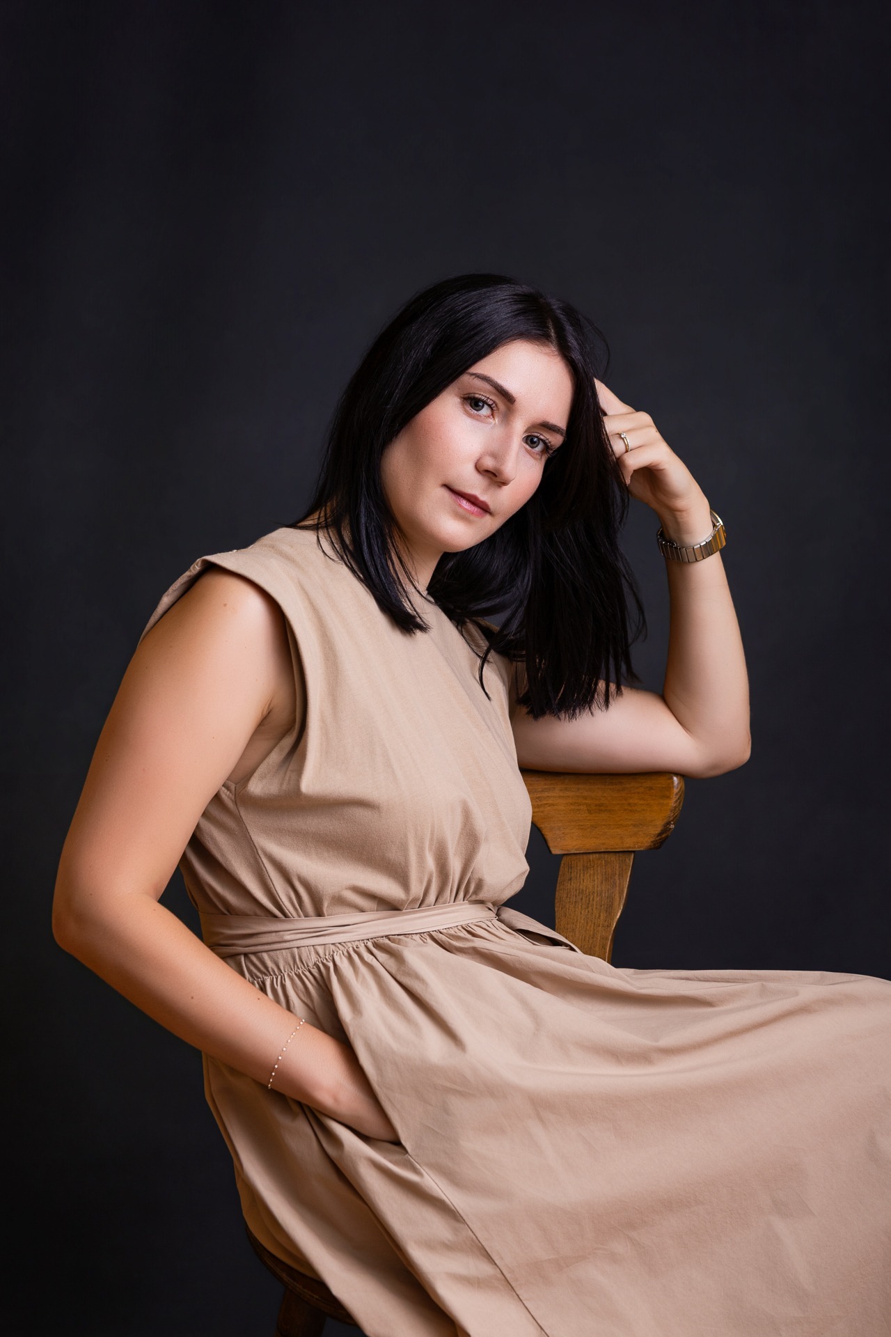 Une femme aux cheveux noirs raides, vêtue d'une robe beige, est assise sur une chaise en bois devant un fond sombre ; elle pose son coude sur le dossier de la chaise et regarde l'objectif — photo prise par un photographe de mariage près de Langres.