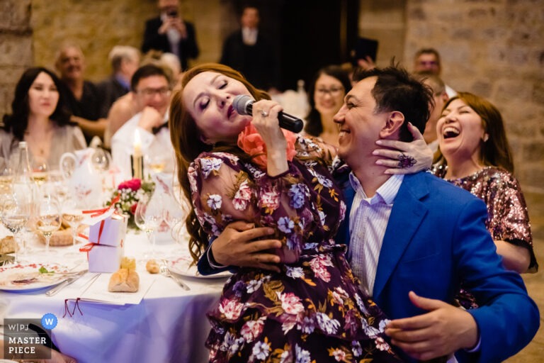 Une invitée chante au micro pendant le dîner, assise sur les genoux d'un invité qui rit.
